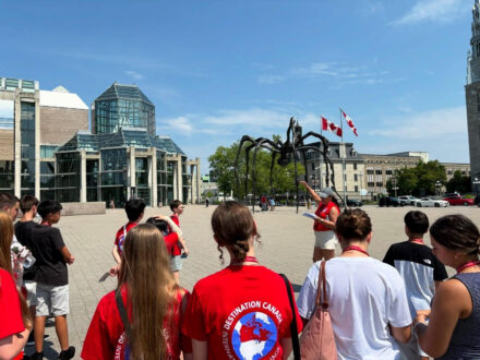 Un groupe d’étudiants en excursion, portant des t-shirts rouges, écoute un guide devant une grande sculpture d’araignée dans une place ouverte, entourée de bâtiments modernes et de drapeaux canadiens flottant au vent journée très ensoleillée.